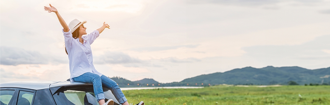Woman sitting on a car with arms raised in celebration, looking at beautiful mountain scenery.