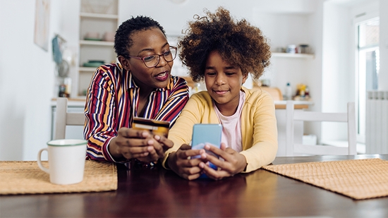 Una madre y su hija se sientan en una mesa mirando un smartphone, sosteniendo una tarjeta de crédito.