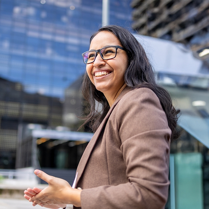 A smiling woman with dark hair and glasses wearing a brown blazer stands outdoors with a modern glass and steel office building in the background.
