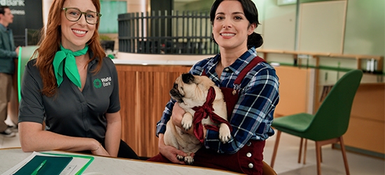 A WaFd Bank employee and a woman holding a pug sit together at a table inside a bank branch.