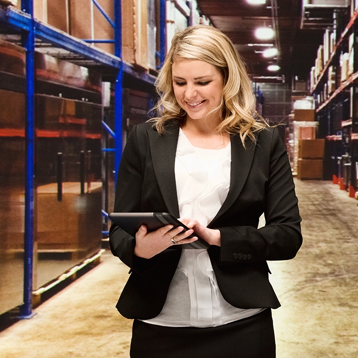 A smiling Blonde woman in a business suit is standing in a warehouse aisle with tall metal shelving filled with cardboard boxes, looking down at and operating a digital tablet.
