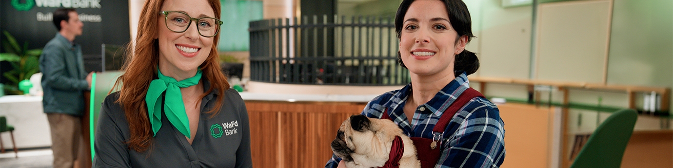 A WaFd Bank employee and a woman holding a pug sit together at a table inside a bank branch.