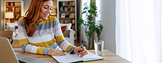 Woman seated at a dining table, planning her time with a notebook and laptop.