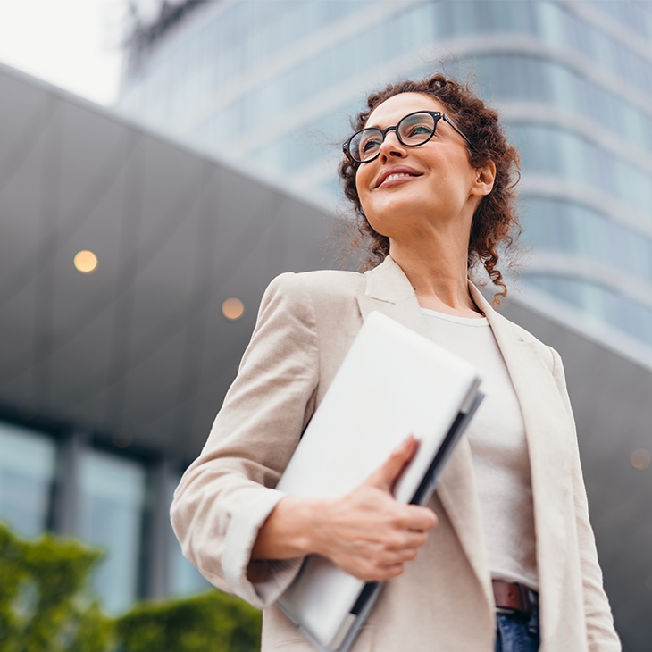 A confident, smiling businesswoman in a light blazer and glasses stands outside a modern building, holding a laptop.