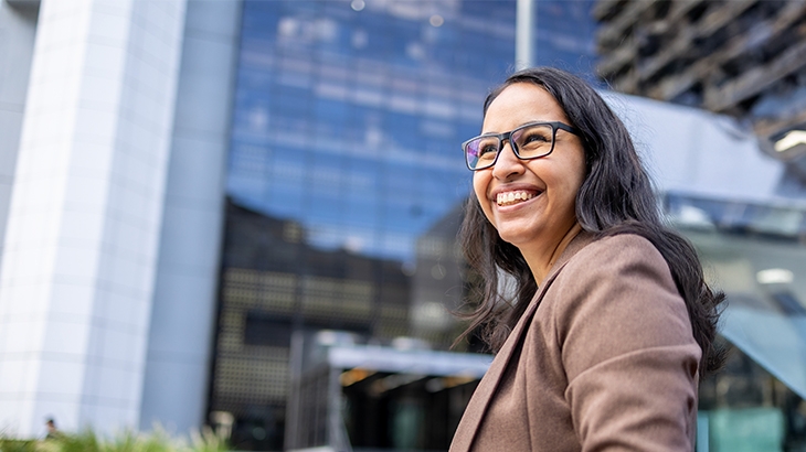 A smiling woman with dark hair and glasses wearing a brown blazer stands outdoors with a modern glass and steel office building in the background.