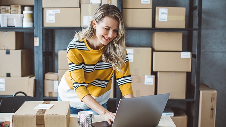 Woman in a yellow and black striped sweater works on a laptop surrounded by cardboard shipping boxes in a small business or warehouse.