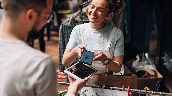A vendor smiles while accepting a contactless mobile payment from a customer using a smartphone and card reader.