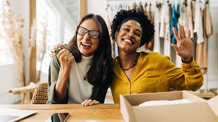 A diverse pair of happy businesswomen, smiling and gesturing enthusiastically in their clothing shop.