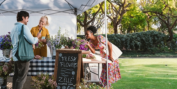 People buying flowers at an outdoor booth with a Zelle accepted here sign.