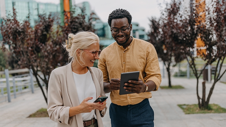 Two smiling professionals standing outdoors, looking at a tablet together.