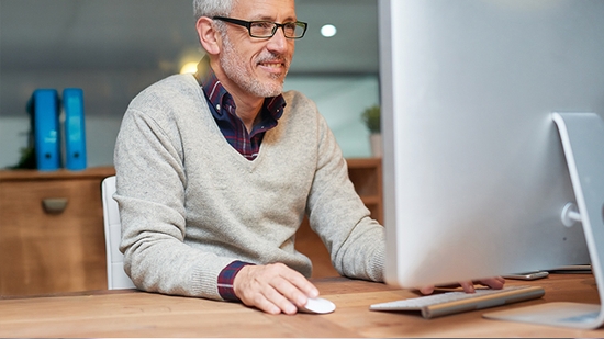 Smiling, middle-aged man with glasses working on a desktop computer in an office.