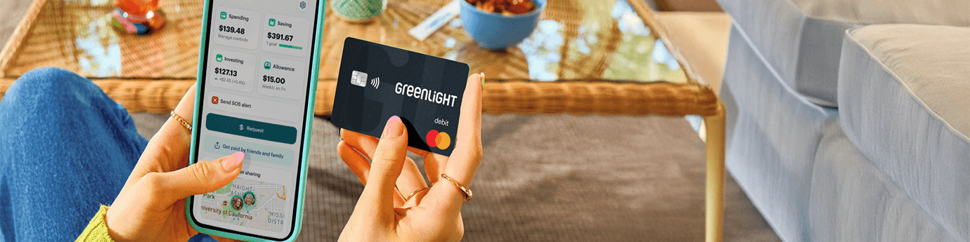 A person sitting indoors holds a smartphone in one hand displaying the Greenlight app with spending, saving, and investing balances, and a black Greenlight debit card in the other. A coffee table with snacks, books, and a drink is visible in the background, creating a casual home setting.