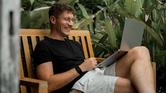 A man sitting on a bench, smiling while looking at his laptop.