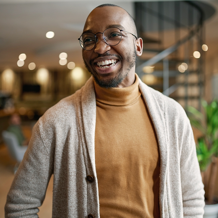A smiling Black man wearing glasses, a tan turtleneck, and a light-colored cardigan stands in a modern, well-lit office.