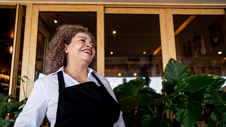 A smiling woman in a white shirt and black apron stands in front of a shop's glass door, surrounded by green plants.