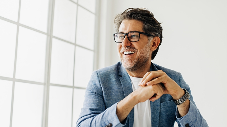 A smiling, middle-aged man with graying hair and a beard, wearing glasses and a light blue blazer, sits in front of a large window.
