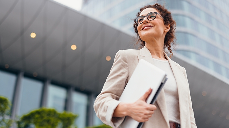 A confident, smiling businesswoman in a light blazer and glasses stands outside a modern building, holding a laptop.