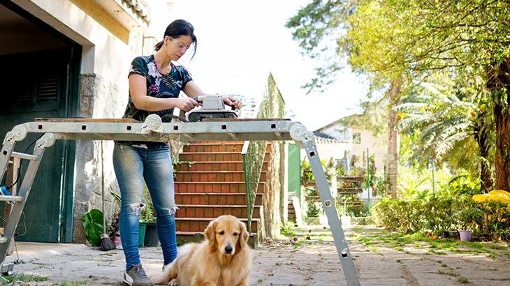 Woman working on a piece of wood outside, dog laying down nearby.