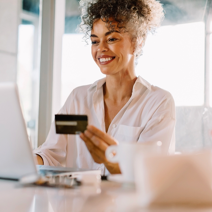 A smiling woman holds a WaFd Bank credit card while working on a laptop in a bright indoor office.