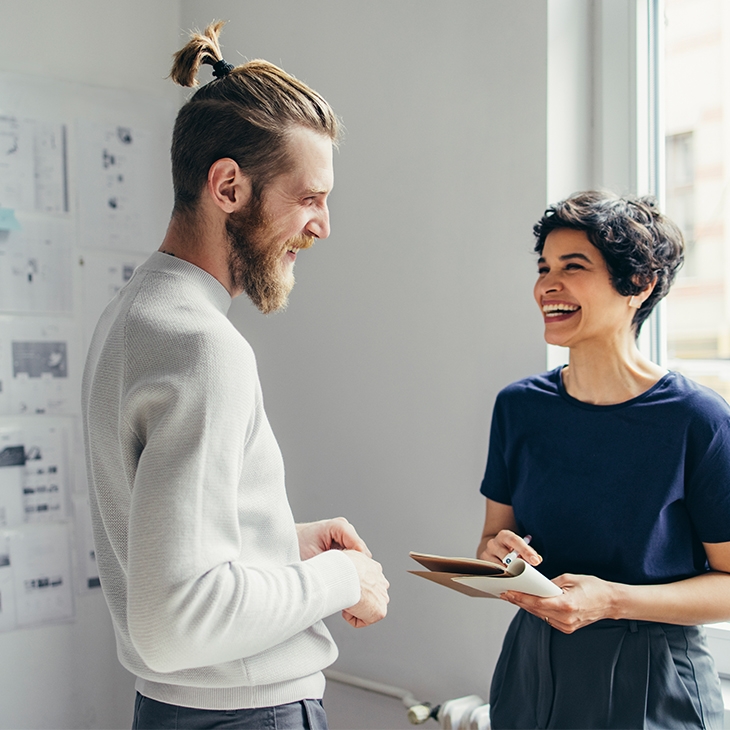 Two smiling colleagues discussing work with documents on a wall in the background.