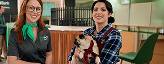 A WaFd Bank employee and a woman holding a pug sit together at a table inside a bank branch.