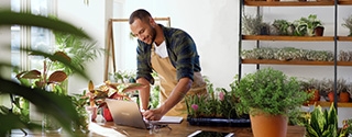 A business owner in an apron working on a laptop at a wooden table in a plant-filled shop or nursery.