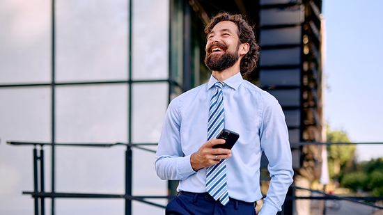 A bearded man in a light blue shirt and striped tie is standing outdoors in front of a modern glass building, laughing while holding a smartphone.