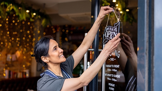 A woman in an apron smiling as she hangs a "Come In, We Are Open" sign on the glass door of a shop.