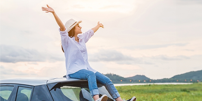 Woman sitting on a car with arms raised in celebration, looking at beautiful mountain scenery.