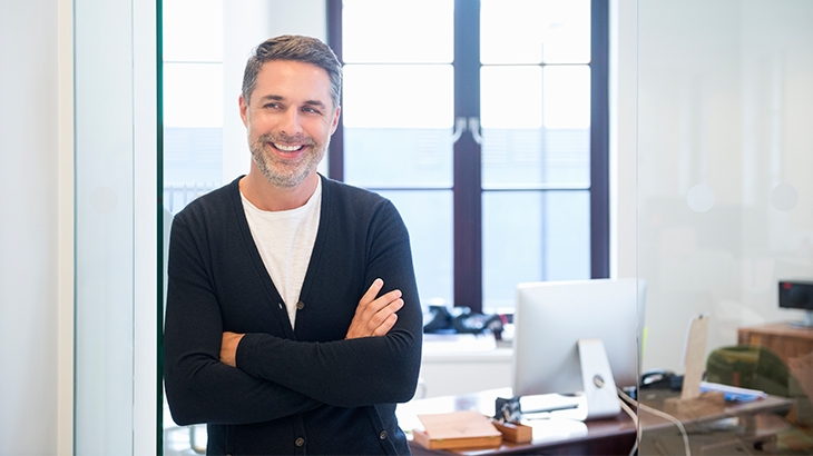 A casually dressed, smiling professional man with salt-and-pepper hair stands with his arms crossed in a modern office.