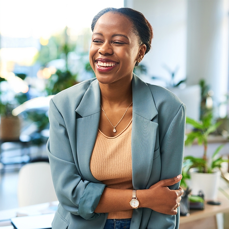 A professional Black woman in a blue blazer and peach top stands smiling in a bright, modern office surrounded by plants.
