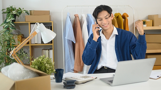 A young man sits at a desk with a laptop and phone, smiling and gesturing with his hand while on a call.
