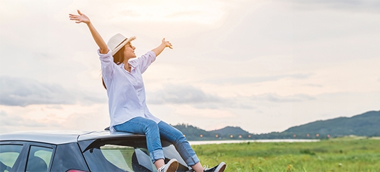 Woman sitting on a car with arms raised in celebration, looking at beautiful mountain scenery.