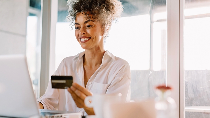 A smiling woman holds a WaFd Bank credit card while working on a laptop in a bright indoor office.