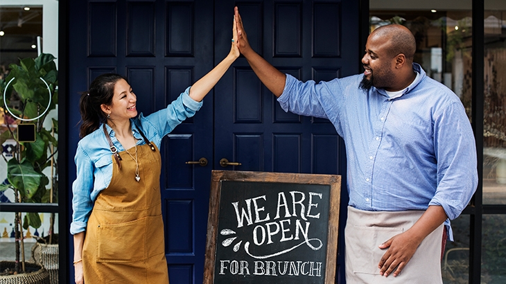 New restaurant owners high-five over a 'we are open' sign.