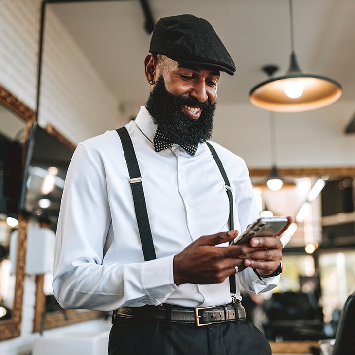 A smiling, well-dressed man with a full beard, wearing a newsboy cap, bow tie, and suspenders, looks at his mobile phone in what appears to be a barbershop or retail space.