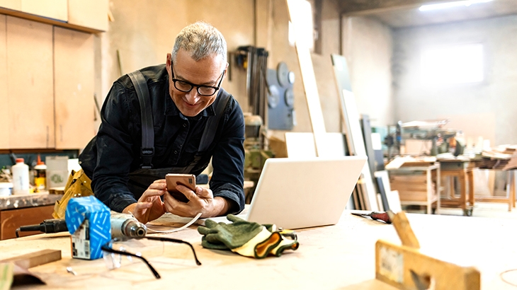 A man in a workshop wearing glasses and overalls smiles while looking at his smartphone, leaning over a workbench with a laptop, tools, and safety gear.