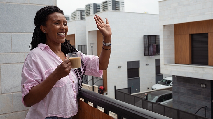 A smiling woman with braided hair stands on a balcony, holding a coffee cup and waving, with modern white townhouses in the background.