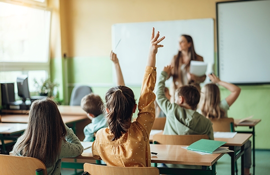 Student raising hand in classroom with other students and teacher
