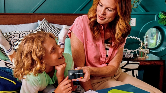 A mother sitting in a bedroom hands a Greenlight debit card to her young child.