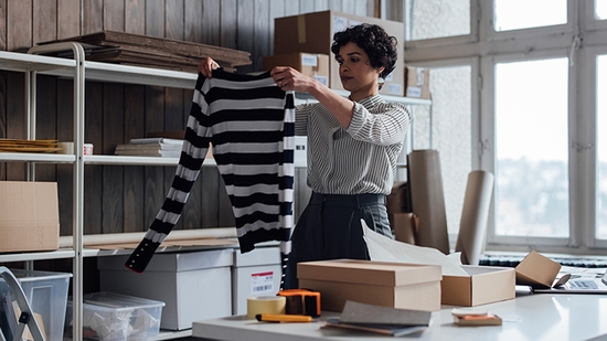 A woman in a warehouse setting holds up a black and white striped sweater, preparing it for packaging.