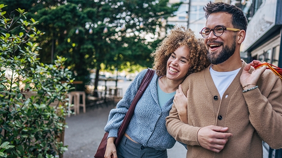 A happy couple walking together outdoors, with the woman leaning her head on the man's shoulder as he carries shopping bags.