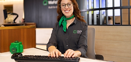A smiling WaFd Bank representative in a branded shirt and green scarf sits at a desk, providing professional support to business clients.