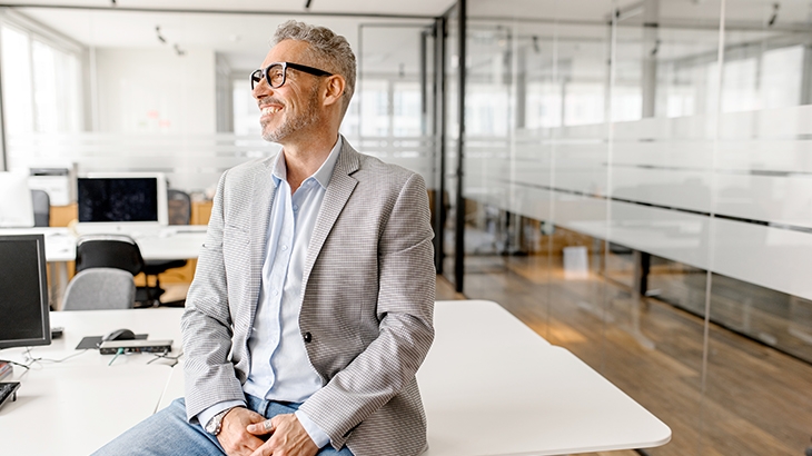 A smiling, gray-haired businessman in a light gray blazer and jeans sits on a desk in a modern office, looking off to the side.