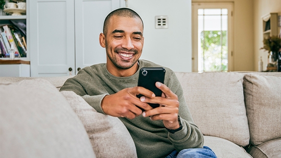 A smiling man relaxes on a living room couch while using a mobile phone.