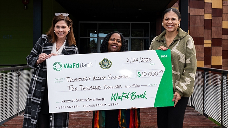 Three women stand outdoors holding a large ceremonial WaFd Bank check for $10,000 made out to the Technology Access Foundation, dated February 24, 2026, and noted as a 'Halfcourt Shooting Contest Winner'.