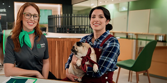 A WaFd Bank employee and a woman holding a pug sit together at a table inside a bank branch.