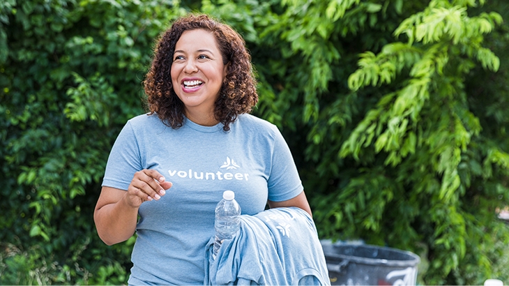 A smiling woman with curly brown hair wears a light blue "Volunteer" t-shirt and stands outdoors, holding a water bottle and a folded shirt.