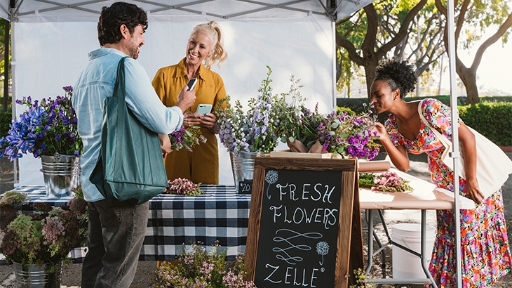 People buying flowers at an outdoor booth with a Zelle accepted here sign.