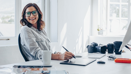 A professional woman wearing glasses and a gray shirt sits at a white desk in a bright office, smiling at the camera while using a stylus on a drawing tablet.